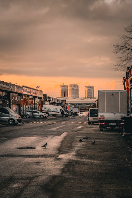 A view of a commercial parking area at sunset, showing a variety of vehicles including a white delivery van, a small silver car, and a larger grey box truck parked near storefronts on either side. The parking lot surface appears wet, with visible puddles and scattered pigeons walking on the pavement. In the background, several modern high-rise buildings are visible against a dramatic sky with grey and orange clouds. Part of the scene features a person walking across the lot, possibly involved in a home relocation or moving activity. The environment suggests an urban setting suitable for loading or unloading furniture, boxes, and packing materials, consistent with house removals and furniture transport operations, such as those provided by Man with Van Harringay.