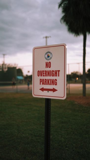 A rectangular white sign with a red border and bold red lettering that reads 'NO OVERNIGHT PARKING,' mounted on a black metal pole. The sign features the Haringey Council emblem at the top and an arrow pointing left and right at the bottom. It is situated outdoors on a grassy area near a paved path or driveway, with a background of a park or sports field, palm trees, and an overcast sky. The scene suggests a residential or urban environment where parking restrictions are in place, relevant to household removals and transport logistics in Haringey, UK, as detailed on the page about parking rules and Harringay removals compliance.
