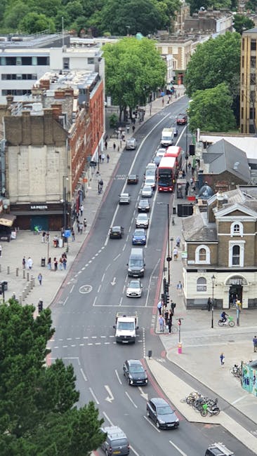 An aerial view of a city street with parked and moving cars, including a red double-decker bus in the distance, bordered by various buildings such as a brick retail space and a small white church-like structure. Tall, leafy green trees line both sides of the road, providing shade. Sidewalks are populated with pedestrians, some walking and others waiting at crossings. Bicycles are parked near a corner, and a few vehicles are seen loading or unloading in a street that appears to be part of a residential and commercial area. The scene is well-lit with daytime natural light, showing typical urban activity consistent with house removal and moving logistics. This image, associated with Haringey Council parking rules and Harringay removals, highlights the busy environment in which furniture transport and packing for home relocation take place, with Man with Van Harringay operating in this area.