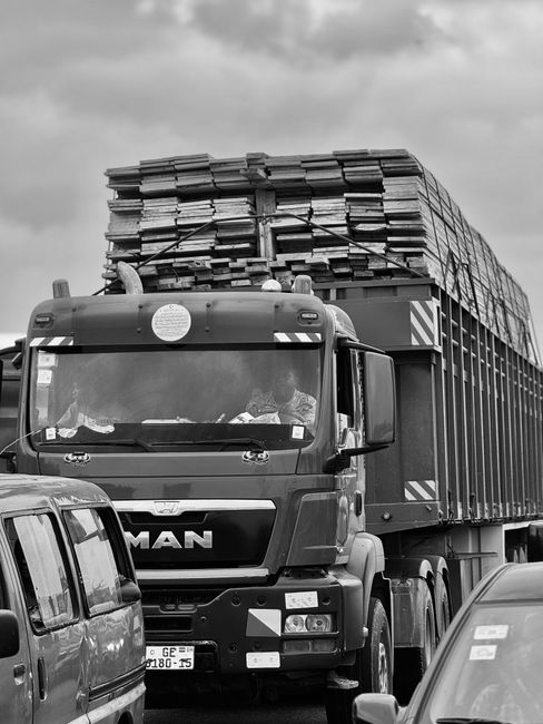 A construction site scene showing a large delivery truck with an open back, filled with stacked bags of building materials. Several workers are involved in the loading process, with some carrying bags