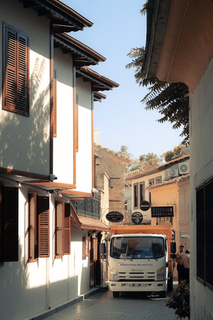 A narrow street in Harringay is shown with two and three-storey residential buildings on either side, featuring white walls and brown wooden window shutters. The street is paved and includes a white flatbed van parked close to one building, with its rear end aligned near the doorway. A person is partially visible beside the van, appearing to be involved in a home relocation process. The area is lit by daylight, and in the background, there are trees and additional buildings, indicating an urban neighbourhood setting. This scene illustrates the typical environment for furniture transport and packing during a house move, with Man with Van Harringay providing moving services in a confined street environment, highlighting the logistical considerations of loading and unloading in such spaces.