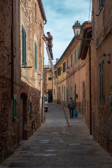 A narrow cobblestone street in a historic residential area, with closely packed old stone and plaster buildings on either side. A man is standing on a tall, extendable ladder leaning against a building, working near the window. The ladder is supported by another person on the ground who appears to be assisting. The street is partially shaded by the buildings, with a traditional street lamp attached to one of the walls. A person walking down the street carries a large bag, heading away from the camera. This scene illustrates a home relocation or repair process, with visible packaging materials and a focus on the narrow access typical for house removals in such environments, as covered by Man with Van Harringay.