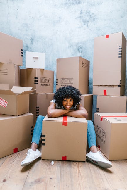 A young woman with curly hair and casual clothing sits on a wooden floor, smiling with her arms resting on a large cardboard moving box in front of her. She is surrounded by numerous stacked cardboard boxes of various sizes, some sealed with red or black tape, and others partially open to reveal packing materials. The background features a textured blue wall, and the scene appears to be inside a room prepared for a home relocation. The presence of the boxes and the casual, relaxed demeanor of the woman suggest she is involved in packing or unpacking during a moving day, which is relevant to house removals and furniture transport services offered by a company like Man with Van Harringay.