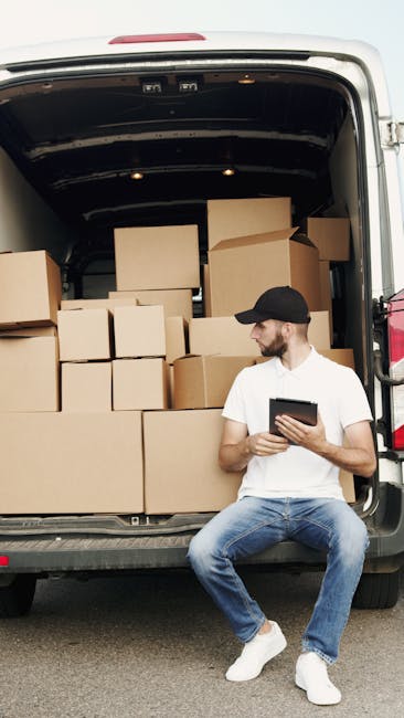 A young man dressed in a white t-shirt, blue jeans, and a black cap is seated on the ground beside an open cargo van during a home relocation. The van's interior is filled with stacked cardboard boxes of various sizes, some partially covered with plastic wrapping materials. Inside the van, the boxes are arranged in multiple layers to optimize space for furniture transport and packing during the moving process. The man is holding a tablet, possibly coordinating or checking an inventory list, as he looks towards the side. The scene takes place outdoors under natural daylight, with the rear doors of the van open to reveal the inside. The setting suggests an element of the loading process where packing materials and boxes are being organized for transportation, indicative of professional removals by companies such as Man with Van Harringay, supporting house removals and moving services.