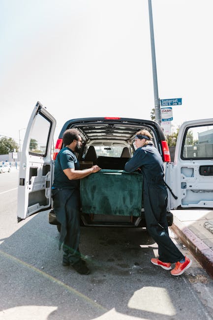 Two professional movers from Man with Van Harringay are loading a large, green upholstered piece of furniture, possibly a footstool or small sofa, into the back of a white panel van parked on a city street. The movers are dressed in dark work uniforms, with one wearing a face mask and the other wearing safety glasses, as they carefully lift and position the furniture inside the vehicle. The van's rear doors are wide open, revealing an interior prepared for furniture transport, with a focus on securing items during home relocation. The scene is outdoors on a paved pavement near a lamppost and street sign, with clear daylight and a blue sky overhead. This image illustrates the process of packing and loading furniture as part of professional removal services, aligned with the services offered by Man with Van Harringay, highlighting the handling of household items during a home move or relocation.