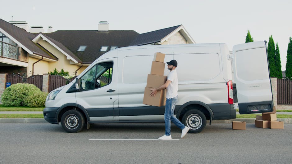 A young man dressed in a white t-shirt, blue jeans, and a black cap is seated on the ground beside an open cargo van during a home relocation. The van's interior is filled with stacked cardboard boxes of various sizes, some partially covered with plastic wrapping materials. Inside the van, the boxes are arranged in multiple layers to optimize space for furniture transport and packing during the moving process. The man is holding a tablet, possibly coordinating or checking an inventory list, as he looks towards the side. The scene takes place outdoors under natural daylight, with the rear doors of the van open to reveal the inside. The setting suggests an element of the loading process where packing materials and boxes are being organized for transportation, indicative of professional removals by companies such as Man with Van Harringay, supporting house removals and moving services.