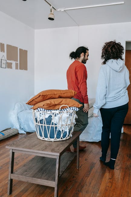Inside a residential property during a home relocation, two individuals are seen near a doorway, engaging in the packing and moving process. One person, dressed in a red top and dark pants, is standing and facing a white wall, while the other, wearing a light grey hoodie and dark jeans, is walking away. In the foreground, a wooden table holds a laundry basket filled with wrapped household items and three large orange cushions. Against the wall, there are partially packed boxes, some wrapped in plastic or fabric, and a brown sofa with protective covers. The room features wooden flooring, a white ceiling with a ceiling-mounted light fixture, and four cardboard panels on the wall possibly indicating ongoing renovation or preparation for moving. The environment reflects the logistics involved in furniture transport, packing, and organizing belongings during a house removal, with Man with Van Harringay providing professional moving services.