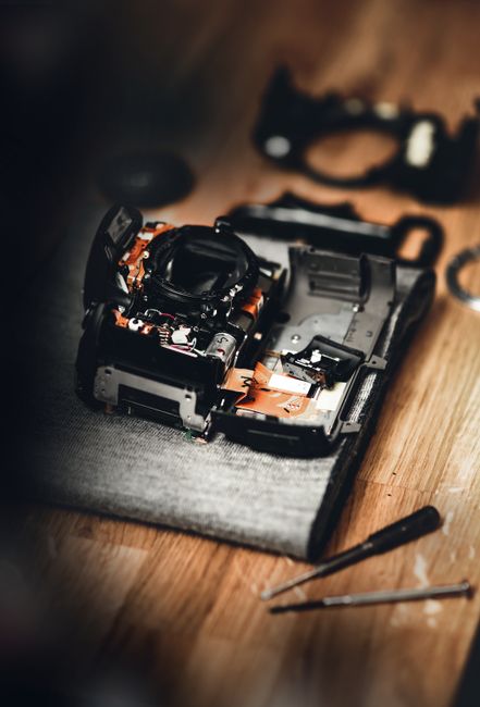 A disassembled digital camera with its internal components exposed, placed on a textured grey surface on a wooden floor. Surrounding the camera are various tools including a screwdriver, a pair of tweezers, and small screws scattered nearby, indicating an ongoing repair or maintenance process. In the background, there is a partially visible black camera strap and a black object that appears to be part of the camera's body or accessories, all set in a well-lit environment. This scene illustrates the detailed work involved in repairing or preparing equipment for a house or furniture move, consistent with professional removals and packing services offered by Man with Van Harringay, as part of a home relocation process.