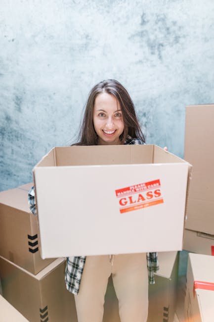 A young woman with long brown hair, smiling and holding an empty cardboard box labeled 'FRAGILE GLASS' in red text, standing in a room with a light blue, textured wall. She is surrounded by several ot