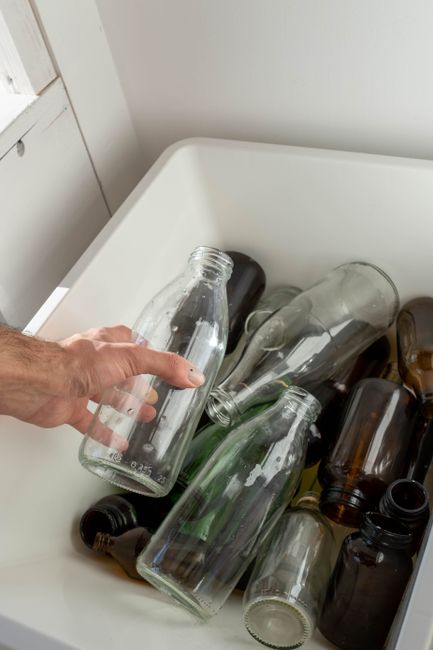 A person's hand holding a clear glass bottle over a white plastic container filled with several other empty glass bottles and jars, some of which are brown or green. The jars are of various sizes and
