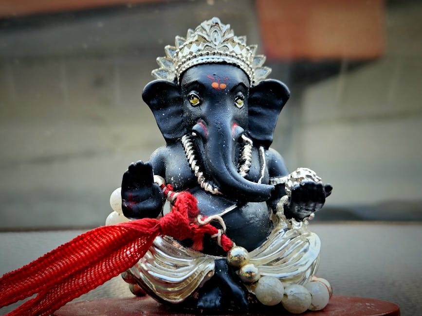 A detailed close-up image of a black Ganesha statue, a Hindu deity with an elephant head, seated on a surface. The statue features intricate silver crowns and ornaments, with a red cloth tied around its trunk and a string of white beads draped over its shoulders. The statue's large ears are prominent, and it has small, attentive eyes with decorative red markings on its forehead. The background includes a blurred wall and a window or door frame, suggesting the statue is placed indoors or in a shaded area, with soft lighting highlighting the detailed craftsmanship. The craftsmanship and materials indicate it is a decorative piece, possibly used for religious or cultural purposes. This image can be associated with themes of home decoration, cultural collection, or spiritual items, relevant to moving or packing services by companies like Man with Van Harringay involved in household relocations.