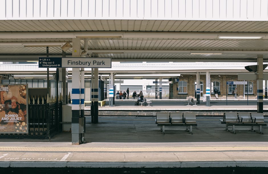 Finsbury Park railway station platform with a covered waiting area featuring multiple metal benches and a sign indicating the station’s name. The platform has a concrete surface with tactile paving along the edge for safety. In the background, additional platforms and waiting passengers can be seen, some sitting on benches or standing near the edge. A poster advertising 'Romeo and Juliet' is mounted on a fence near the platform. Overhead, the station’s roof is supported by metal columns, and there's a clear sky visible beyond the station infrastructure. The scene depicts a typical station environment suitable for house removals or transit-related activities, consistent with the services offered by Man with Van Harringay, including furniture transport and home relocation logistics.